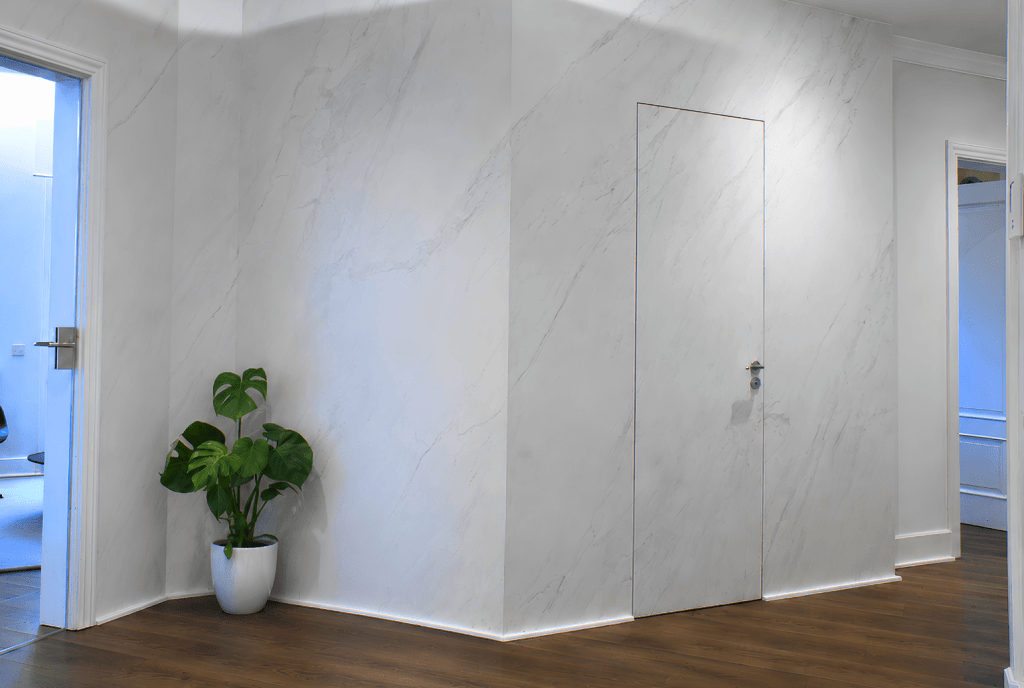 Pasillo con paredes de mármol y una puerta oculta, decorado con una planta verde en maceta blanca.